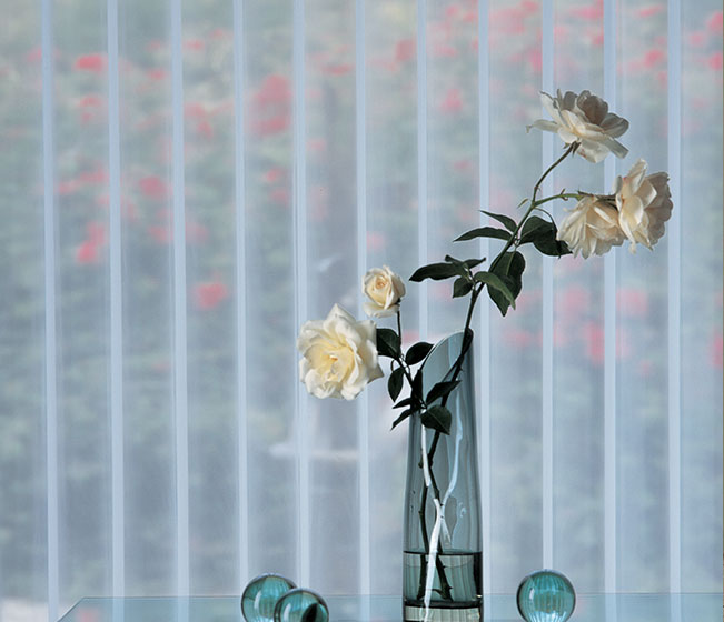 A vase with white roses in front of vertical sheer blinds.
